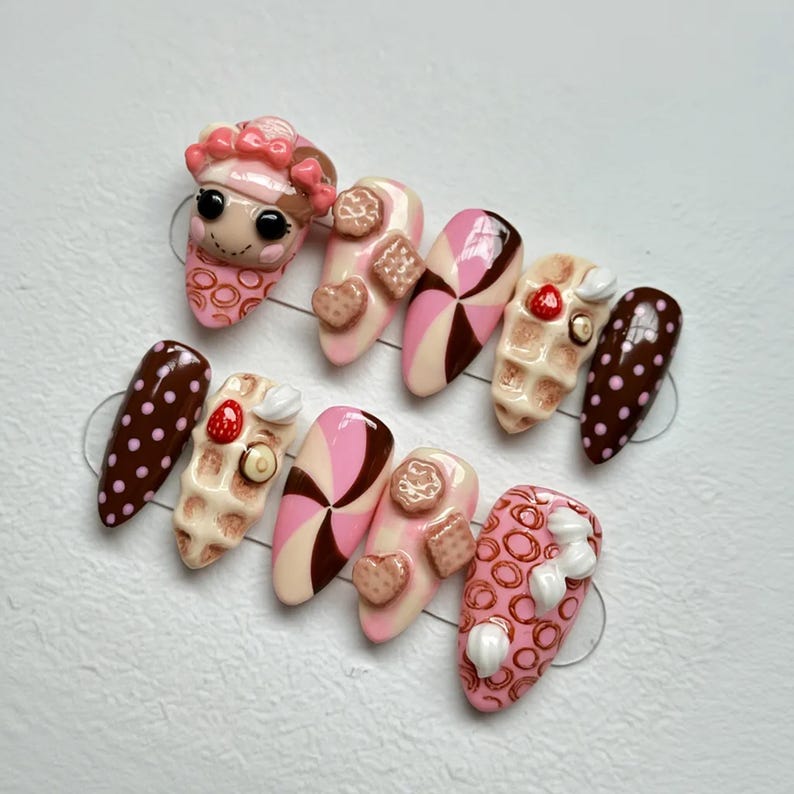 May include: A set of ten almond-shaped press-on nails with a dessert theme. Designs include pink and brown swirls, waffle and cookie accents, and a nail with a pink face and a hat. Some nails have a brown polka dot pattern.