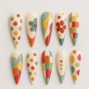 May include: A set of ten almond-shaped press-on nails featuring a cream base color with various hand-painted designs including multicolored polka dots, floral motifs, wavy lines, and geometric grid patterns accented with thin gold metallic lines.