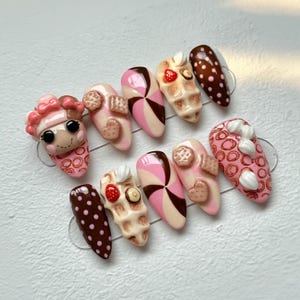 May include: A set of ten almond-shaped press-on nails with a dessert theme. Designs include a doll face, waffles, hearts, and polka dots in shades of pink, brown, and cream. Some nails feature 3D embellishments.
