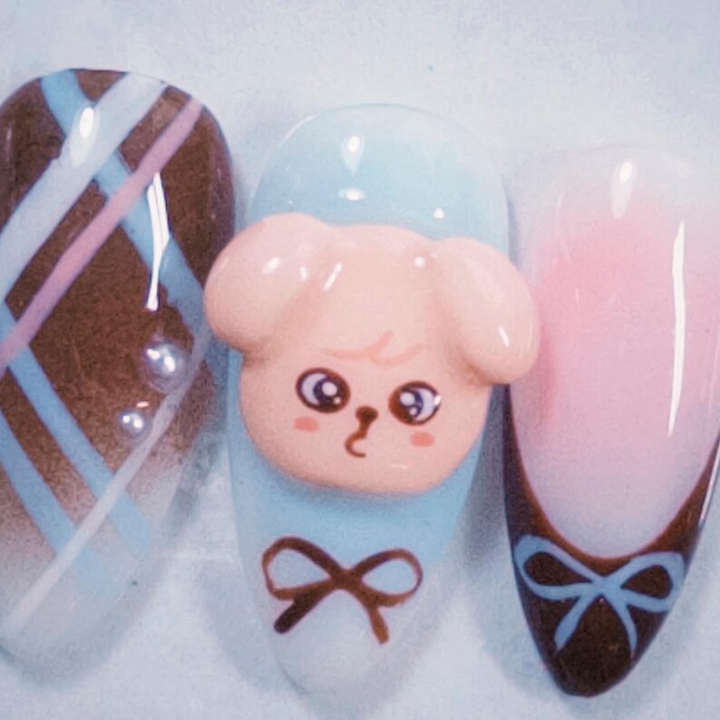 May include: A set of three manicured nails. One nail features a brown and white plaid design with a pearl accent. Another nail has a light blue base with a 3D pink puppy face and a brown bow. The third nail has a pink and white ombre with a brown and blue bow.