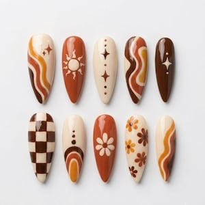 May include: A set of ten almond-shaped press-on nails featuring a warm color palette of cream, orange, and dark brown. Designs include wavy lines, a sun motif, stars, a checkerboard pattern, concentric arches, and small daisy-like flowers.