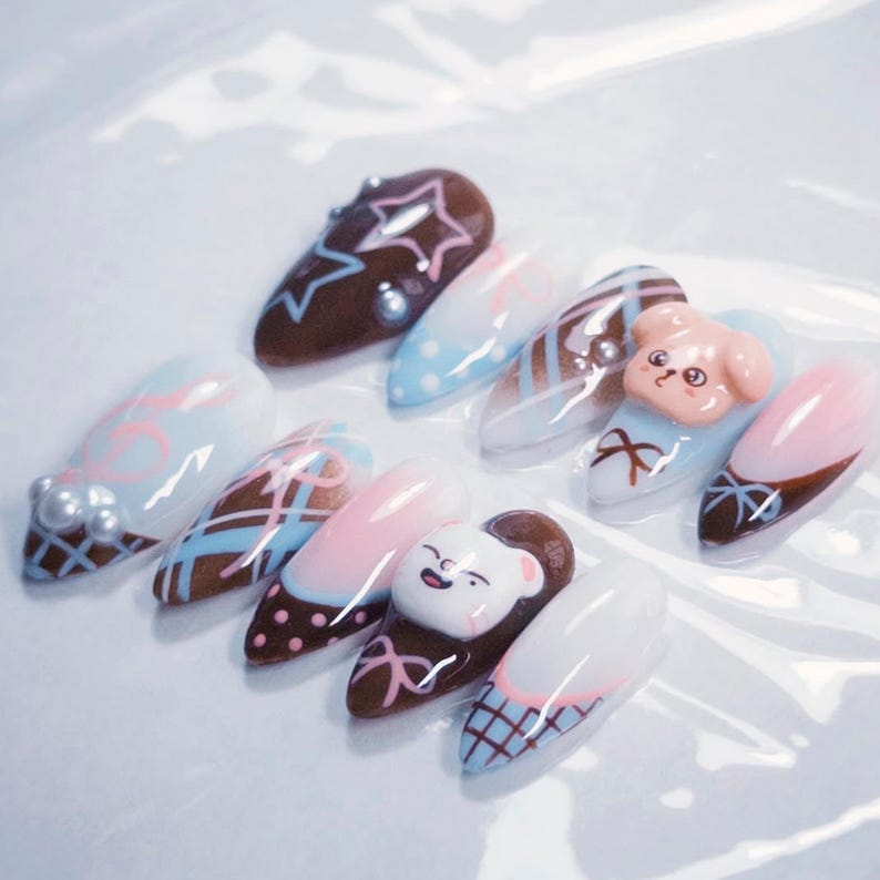 May include: A collection of almond-shaped, decorated fingernails. Designs include a brown nail with a star, a light blue nail with a bow, and a pink nail with a smiling bear. Other nails feature pearl accents and geometric patterns.