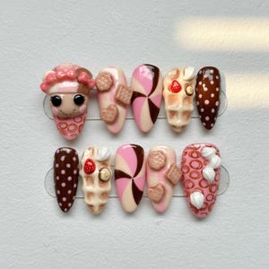 May include: A set of ten almond-shaped false nails with a dessert theme. Designs include pink, brown, and white colors, featuring waffle, cookie, and swirl patterns. Some nails have 3D embellishments.
