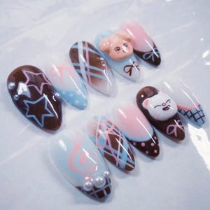 May include: A collection of almond-shaped artificial nails with various designs. Some nails feature brown star patterns, while others have pink and blue plaid or polka dot patterns. Several nails have 3D animal figures and pearl accents.