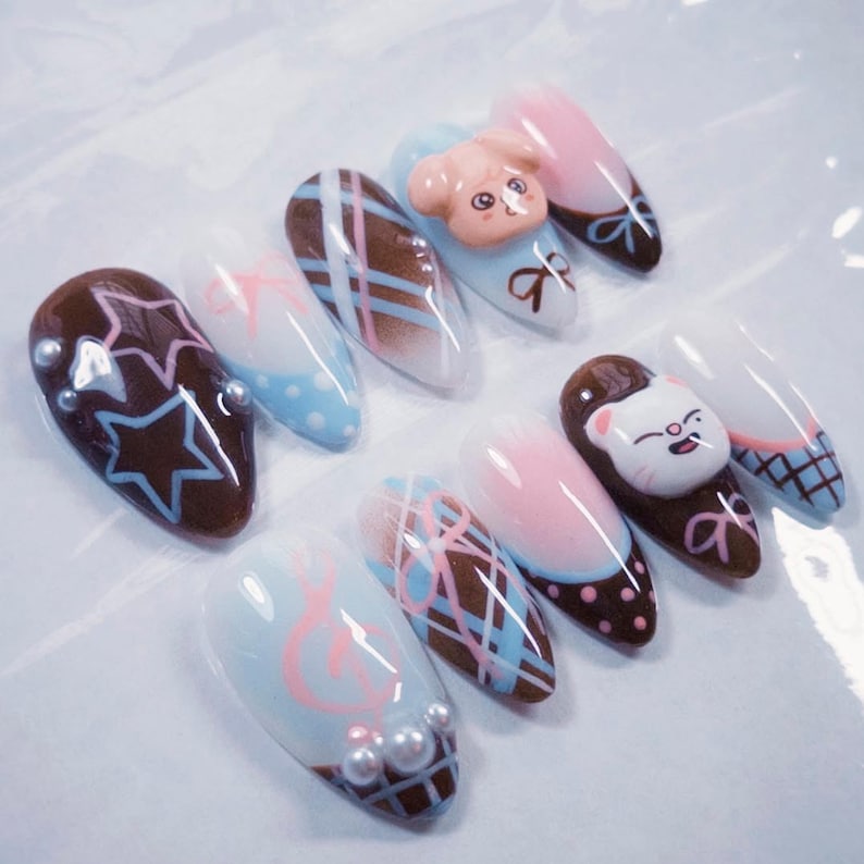 May include: A collection of almond-shaped artificial nails with various designs. Some nails feature brown star patterns, while others have pink and blue plaid or polka dot patterns. Several nails have 3D animal figures and pearl accents.