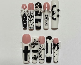 May include: A set of press-on nails with various designs. The nails feature black and white patterns, bows, crosses, and the word "Angel". Some nails have a pink base, while others are white or black. The nails are displayed on clear plastic holders.