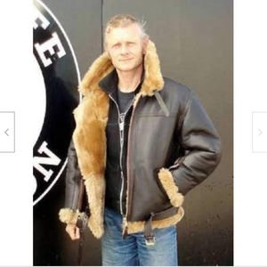 Op de afbeelding: Een donkerbruine leren jas met een beige schapenvachtkraag en voering. De jas heeft een ritssluiting, een gespriem in de taille en schapenvachtmanchetten. De persoon draagt een blauwe spijkerbroek.