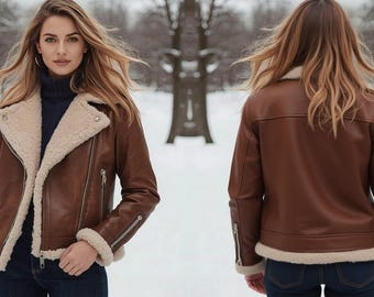 May include: A brown leather jacket with a cream-colored shearling lining and trim. The jacket has a classic biker style with a notched collar, asymmetrical zipper, and zippered cuffs. The model is wearing the jacket in a snowy outdoor setting.