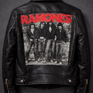 Puede incluir: Chaqueta de cuero negro con un gráfico de la banda Ramones en la espalda. El gráfico es en blanco y negro con el nombre de la banda en rojo. La chaqueta tiene un estilo motero clásico con cuello de solapa y bolsillos con cremallera.