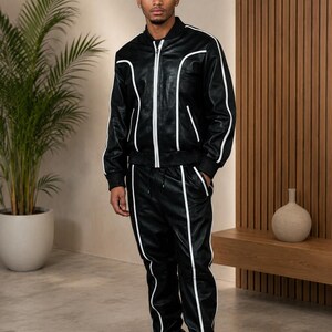 Peut inclure: Ensemble de surv&ecirc;tement en cuir noir avec passepoil blanc. La veste est zipp&eacute;e &agrave; l'avant et le pantalon a une taille &agrave; cordon. La tenue est port&eacute;e avec des bottes noires. Le mod&egrave;le est dans une pi&egrave;ce moderne aux tons neutres.