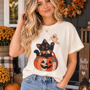 May include: A cream-colored t-shirt featuring a graphic of a black cat sitting inside a patchwork pumpkin, with a small ghost floating above. The cat has a colorful plaid collar, and the pumpkin is decorated with various orange and red patterns.