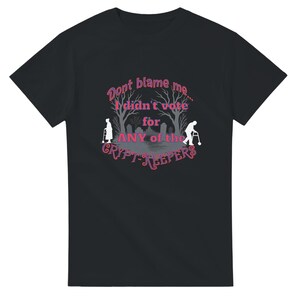 May include: A black short-sleeve t-shirt featuring a graphic of two elderly figures with walkers in a graveyard. The text reads: "Dont blame me... I didn't vote for ANY of the CRYPT-KEEPERS" in pink and purple lettering.