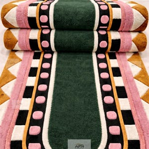 May include: A rectangular rug with rounded ends, featuring a central dark green panel surrounded by concentric borders of black, pink circles, white squares, and mustard yellow. The design includes a geometric zigzag pattern at the edges.