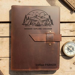 May include: A brown leather journal featuring a dark engraved graphic of a tent, campfire, mountains, and trees with the text WANDER EXPLORE DISCOVER. The cover includes a leather strap closure with two brass rivets and the name William PARKER.