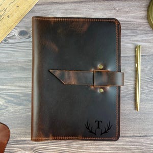 May include: A dark brown leather journal cover featuring a horizontal strap closure secured with two brass-colored rivets. The bottom right corner displays a black engraved letter T centered between two stylized deer antlers. A gold pen lies nearby.