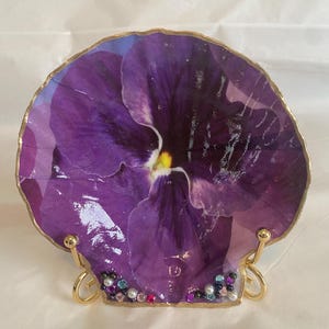 May include: A decorative, shell-shaped dish featuring a vibrant purple pansy flower design. The dish has a gold-colored rim and is supported by a gold-colored stand. The base is adorned with small, colorful beads.