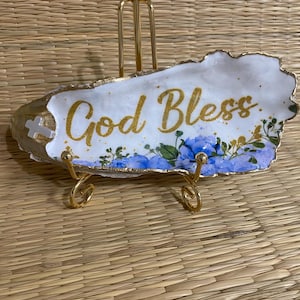 May include: A decorative, white, oblong dish with gold trim and a gold stand. The dish features the gold text "God Bless" with blue floral accents. A small cross is on the left side of the dish.