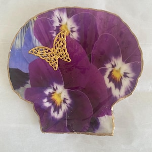 May include: A decorative coaster featuring a vibrant floral design with purple pansies and a gold-colored butterfly accent. The coaster has a gold-colored edge and is shaped like a seashell.