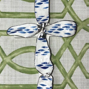 May include: A decorative cross made from white, irregularly shaped pieces with a blue fish scale pattern. The pieces are connected with silver-colored metal. The cross is set against a green and white trellis patterned background.