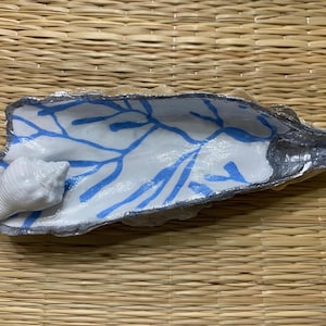May include: A decorative seashell dish with a white interior featuring a blue branch-like pattern. A small white seashell rests inside the dish. The dish has a silver-coloured rim and is set on a woven mat.