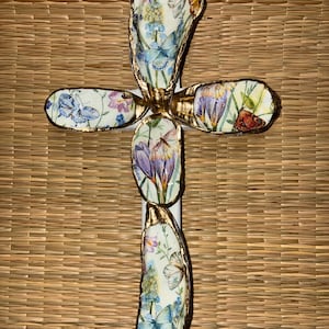 Peut inclure: Une croix décorative composée de cinq coquilles d'huîtres individuelles disposées en forme de croix. Chaque coquille présente un motif floral et papillon en découpage dans des tons de bleu, violet et orange, avec des bords dorés.