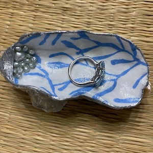 May include: A handmade ceramic jewellery dish with a white and blue leaf pattern. The dish has a silver-coloured rim and contains a silver ring with blue gemstones and small, pearl-like beads.