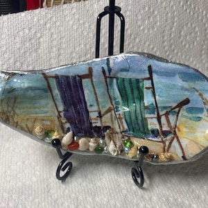 May include: A decorative oyster shell featuring a beach scene with two beach chairs and towels. The shell is painted with blue, green, and purple hues, and is adorned with small seashells and beads. It is displayed on a black metal stand.