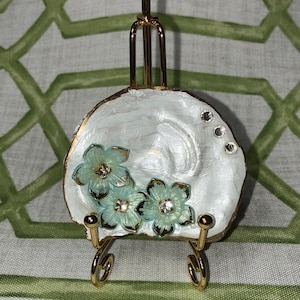 May include: A decorative miniature easel featuring a white, shell-shaped surface. The shell is decorated with three light blue flower embellishments with crystal centres and gold trim. The easel has a gold-coloured frame and legs.