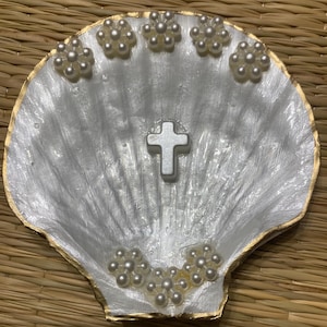May include: A decorative, white seashell-shaped dish with a gold-painted rim. The dish features a silver cross in the centre and is adorned with clusters of faux pearls. The background is a woven, natural-toned mat.
