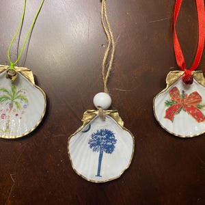 Może przedstawiać: Trzy muszelkowe ozdoby ze złotymi krawędziami. Jedna przedstawia palmę z ozdobami, druga niebieskie drzewo, a ostatnia czerwoną kokardę. Każda ma inny sznurek do zawieszenia: zielony, sznurek i czerwoną wstążkę.