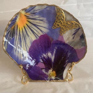 May include: A decorative, shell-shaped dish featuring a vibrant floral design with purple, blue, and yellow hues. A gold-colored, filigree butterfly adorns the dish, which has a gold-painted rim. The dish is displayed on a gold-colored stand.