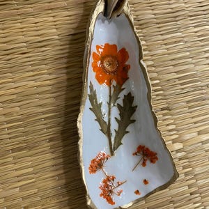 May include: A decorative, oblong dish with a white interior and gold-painted edges. The dish features an orange flower and dried orange floral accents. A seashell is attached to the top of the dish. The dish is on a woven mat.