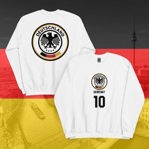 May include: White sweatshirt featuring a circular emblem with a black eagle and the word "DEUTSCHLAND". The back of the sweatshirt displays the same emblem. The front has the emblem and the word "GERMANY" with the number "10".