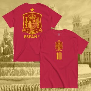 May include: Two red t-shirts with gold emblems and text. One shirt displays the Spanish coat of arms and the word "ESPAÑA". The other shirt has the Spanish coat of arms, the word "ESPAÑA", and the number "10".