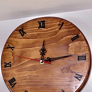 May include: A circular wooden clock with a light brown finish. The clock face has black Roman numerals and black hands, with a red second hand. The clock is mounted on a white wall.