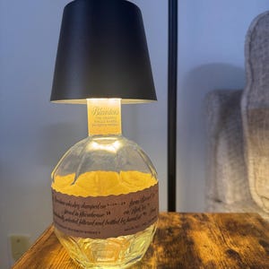 Puede incluir: Una lámpara de mesa única hecha de una botella de whisky bourbon Blanton's reciclada. La lámpara presenta una pantalla negra y una botella de vidrio transparente con una etiqueta. La botella está iluminada con una luz amarilla cálida.