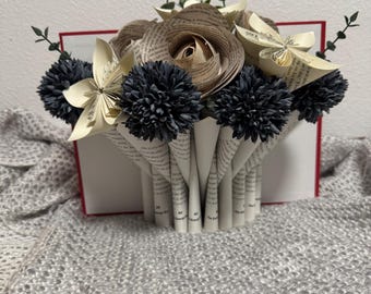 May include: A handcrafted paper flower arrangement. The bouquet features a central rose and other flowers made from book pages, with dark blue pom-pom accents and greenery. The base is a folded paper vase.