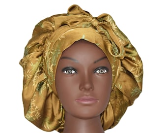 May include: A golden-yellow satin bonnet with a green and white bamboo leaf pattern. The bonnet is tied at the top with a bow and is displayed on a dark-skinned mannequin head. The fabric has a slight sheen.