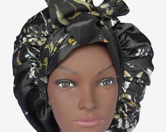 May include: Black satin bonnet with a floral pattern in white, yellow, and orange. The bonnet has a bow on top and is designed to protect hair. The bonnet is on a mannequin head.