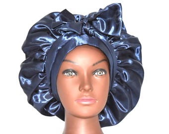 May include: A navy blue satin bonnet with a large bow on top. The bonnet is designed to protect hair and is displayed on a mannequin head. The satin material has a shiny appearance.