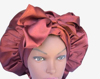 May include: A deep rust-colored satin bonnet featuring a large, structured bow detail on the front. The head covering has a gathered, elasticized edge and a smooth, reflective finish, displayed on a mannequin head to show its shape and volume.
