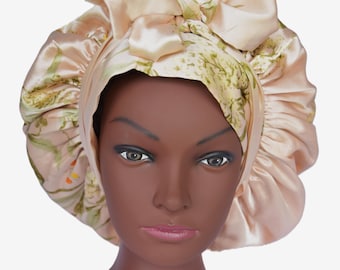 May include: A peach-colored satin bonnet with a large bow on top. The bonnet features a floral pattern in shades of green and yellow. The bonnet is designed to protect hair and maintain hairstyles.