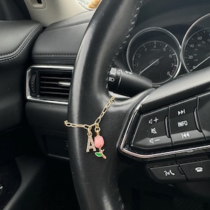 May include: A gold-coloured car charm with a chain, the letter "A" and a pink tulip pendant. The charm hangs from a black steering wheel, with car controls and dashboard visible in the background.
