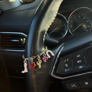 May include: A gold charm bracelet with Western-themed charms hangs from a black steering wheel. Charms include a pink boot, a red star, a cactus, a pink heart, and a pink horseshoe with the word "LUCKY".