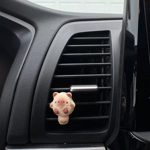 May include: A beige bear-shaped car air vent diffuser with pink details. The diffuser is attached to a car's air vent. The car's interior features black and silver elements.