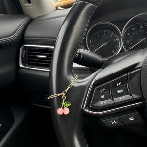 May include: A gold chain charm featuring pink cherries and green leaves hangs from a black steering wheel. The car interior is black with silver detailing. A small soft toy is visible in the background.