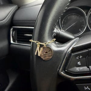 May include: A gold-toned chain charm featuring a rhinestone-encrusted letter N and a circular pendant engraved with a sunburst design and the text YOU ARE MY SUNSHINE, attached to the leather-wrapped rim of a black vehicle steering wheel.