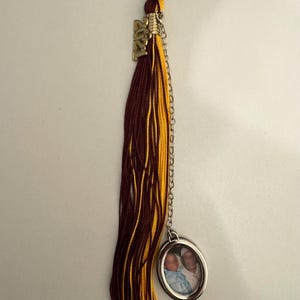 May include: A maroon and gold graduation tassel with a gold-coloured charm reading "2021". A silver-coloured oval pendant with a small photo hangs from a chain attached to the tassel.