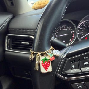 May include: A gold-toned charm bracelet featuring a rectangular white pendant with a red strawberry illustration, a small red cherry charm, a rectangular bar with a clear stone, a heart charm, and a round white pearl-like bead.