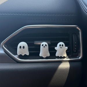 May include: Three small, white, ghost-shaped car air vent clips featuring black painted eyes and mouths. Each ghost has a distinct facial expression, including one with a surprised look and one with a smiling face, attached to the horizontal slats.